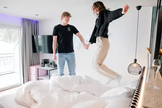 Ein junges Paar springt fröhlich auf einem Bett. Der Mann hält die Hand der Frau, die in der Luft schwebt. Die Szene vermittelt eine entspannte und glückliche Stimmung in einem modernen, hellen Zimmer.