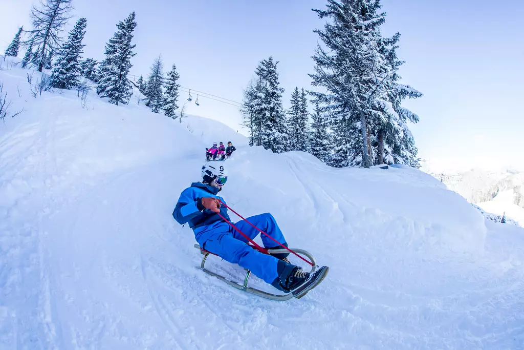 Ein Mensch in blauer Winterkleidung fährt auf einem Schlitten durch verschneite Landschaft. Im Hintergrund sind weitere Personen zu sehen, die ebenfalls Schlitten fahren. Umgeben von hohen, schneebedeckten Bäumen und klaren Himmel zeigt die Szene eine winterliche Freizeitaktivität.