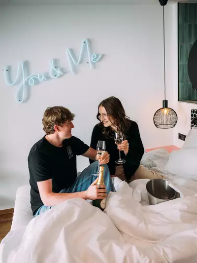 Ein Paar sitzt in einem Bett, umgeben von einer weißen Decke. Sie stoßen mit Gläsern an und lächeln sich an, während eine Flasche Sekt neben ihnen steht. Im Hintergrund leuchtet eine neonfarbene Schrift mit „You & Me“.
