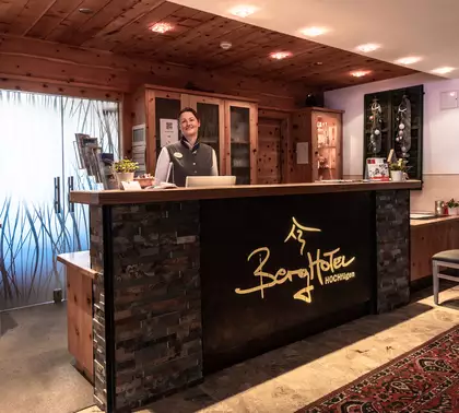 Ein Empfangstresen in einem Hotel mit einer lächelnden Mitarbeiterin. Der Tresen ist aus Stein und Holz, im Hintergrund befindet sich eine durchsichtige Glastür. Regale mit verschiedenen Produkten sind sichtbar, und eine Ecke ist mit Stühlen und Pflanzen dekoriert.