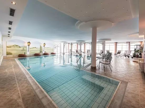 Ein eleganter Innenpool mit klaren Wasser. Der Poolbereich ist von einem hellen Raum umgeben, der mit Fenstern für Tageslicht sorgt. Entspannte Liegen stehen bereit, und dekorative Wände zeigen sanfte Landschaften. Die Atmosphäre ist ruhig und einladend.