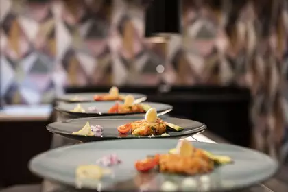 Drei elegante Teller mit kunstvoll angerichtetem Gericht. Jeder Teller enthält eine Mischung aus Gemüse, feinen Kräutern, Garni und einer Zitronenscheibe. Der Hintergrund ist verschwommen, zeigt aber eine moderne und stilvolle Atmosphäre.