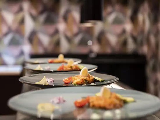 Drei elegante Teller mit kunstvoll angerichtetem Gericht. Jeder Teller enthält eine Mischung aus Gemüse, feinen Kräutern, Garni und einer Zitronenscheibe. Der Hintergrund ist verschwommen, zeigt aber eine moderne und stilvolle Atmosphäre.