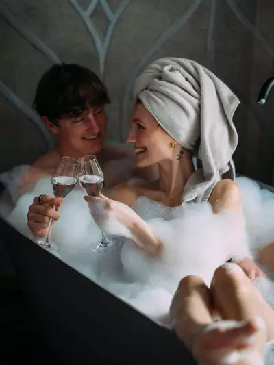 Ein Paar sitzt in einer Badewanne voller Schaum. Sie lächeln sich an und halten zwei Gläser Sektempfang. Die Atmosphäre ist entspannt und romantisch, mit einem Handtuch auf dem Kopf der Frau.
