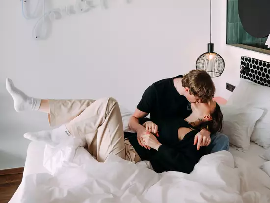 Ein junges Paar liegt auf einem Bett, umarmt sich und küsst sich liebevoll. Die Bettdecke ist zerwühlt und es sind Kissen um sie herum. An der Wand hinter ihnen steht der Schriftzug „You & Me“.