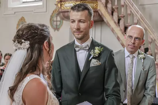 Ein Brautpaar steht sich während der Trauung gegenüber. Die Braut trägt ein elegantes Kleid und einen Schleier, während der Bräutigam in einem Anzug mit Fliege gekleidet ist. Im Hintergrund schaut ein weiterer Mann, sichtbar als Trauzeuge, aufmerksam zu.