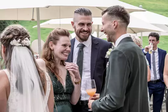 Eine Gruppe von Gästen unterhält sich auf einer Feier im Freien. Zwei Männer lachen miteinander, während eine Frau in einem grünen Kleid lächelt. Im Hintergrund sind weitere Gäste und Sonnenschirme zu sehen. Die Atmosphäre wirkt freundlich und gesellig.