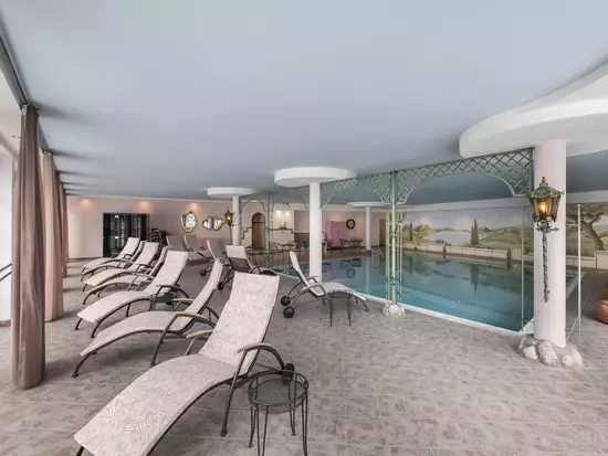 Ein entspannender Wellnessbereich mit mehreren Liegen, einem Schwimmbad im Hintergrund und einem großen Fenster, das Tageslicht hereinlässt. Die Wände sind sanft dekoriert, und es gibt gemütliche Ecken zum Entspannen. Ein ruhiger, einladender Raum für Erholung und Ruhe.