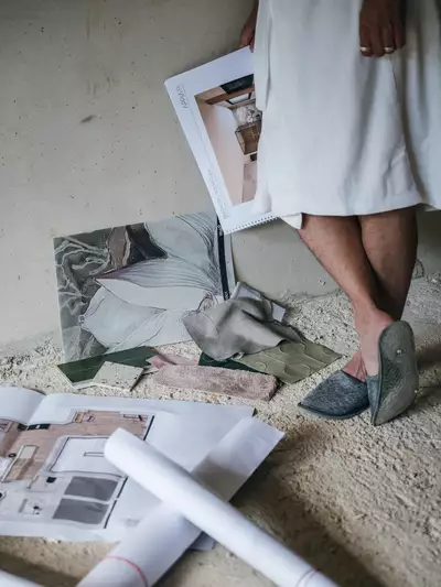Eine Person steht auf einem unebenen Boden und hält eine Mappe mit Innenraum-Design-Entwürfen. Um sie herum liegen verschiedene Stoffmuster und Pläne. Die Kleidung der Person ist hell und sie trägt besondere Schuhe.