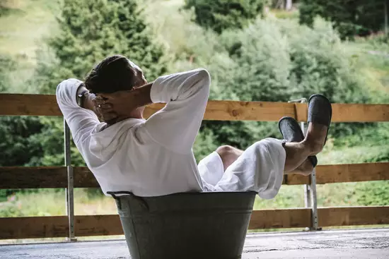 Eine Person sitzt entspannt in einem Eimer auf einem Balkon. Sie trägt einen weißen Bademantel und hat die Füße auf dem Rand des Behälters. Im Hintergrund sind grüne Wälder und Bäume sichtbar, die eine ruhige Naturszene schaffen.