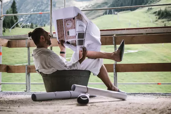 Ein Mann sitzt in einem Eimer und liest eine Bauzeichnung. Er trägt ein weißes Oberteil und hat die Beine lässig angehoben. Im Hintergrund sind grüne Hügel und Berge sichtbar. Rund um ihn liegen einige zusammengerollte Pläne.