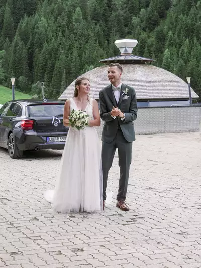 Ein Brautpaar steht lächelnd zusammen, während sie applaudierenden Gästen gegenübertreten. Die Braut trägt ein elegantes, fließendes Kleid und hält einen Blumenstrauß. Im Hintergrund sind grüne Berge und parkende Autos zu sehen. Die Atmosphäre wirkt festlich und jubilierend.