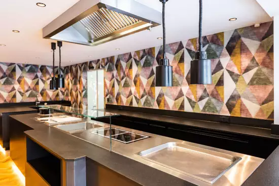 Moderne Küchenansicht mit einer langen, schwarzen Arbeitsplatte. Auf der Arbeitsfläche sind Kochfelder und Edelstahl-Behälter sichtbar. Über der Fläche hängen schwarze Pendelleuchten. Der Hintergrund ist mit einem geometrischen, bunten Muster dekoriert. Langsame Beleuchtung schafft eine einladende Atmosphäre.