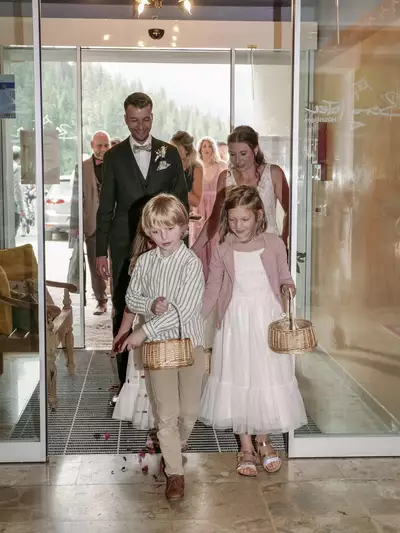 Eine Gruppe von Personen tritt aus einem Gebäude. Im Vordergrund gehen zwei Kinder, ein Junge und ein Mädchen, Hand in Hand und tragen kleine Körbe. Der Junge trägt ein gestreiftes Hemd, die Mädchen ein weißes Kleid. Hinter ihnen folgen mehrere Erwachsene in festlicher Kleidung.