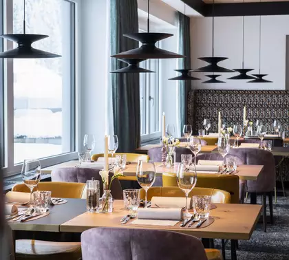 Ein eleganter Restaurantbereich mit mehreren Tischen, die festlich gedeckt sind. Jeder Tisch hat Gläser, Besteck und dekorative Kerzen. Sanfte Beleuchtung von modernen Deckenlampen sorgt für eine einladende Atmosphäre. Große Fenster lassen Tageslicht herein und bieten einen Blick auf die verschneite Landschaft.
