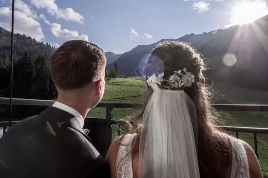 Ein Brautpaar steht auf einem Balkon und blickt auf eine malerische Berglandschaft. Die Frau trägt ein Brautkleid mit einem langen Schleier und Blumen im Haar. Die Sonne scheint hinter den Bergen und erhellt die Szene.