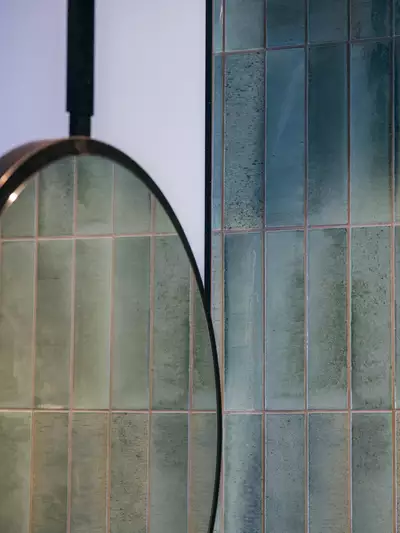 Ein runder Spiegel hängt an einer Wand, die mit modernen, rechteckigen, grünlichen Fliesen verkleidet ist. Die Fliesen zeigen verschiedene Farbtöne und eine glatte Oberfläche, was dem Raum einen eleganten Look verleiht.