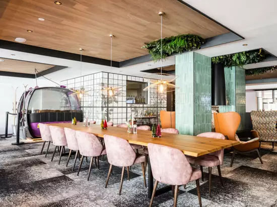 Ein modern gestalteter Restaurantbereich mit einem langen Holztisch und gepolsterten Stühlen in sanften Farben. Im Hintergrund steht eine auffällige, lila Seilbahn. Die Wände sind dekoriert und der Raum verfügt über eine angenehme Beleuchtung und grünes Pflanzenwerk.