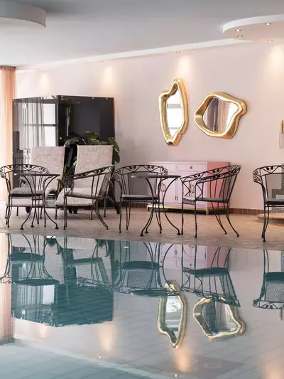 Ein stilvoller Innenraum mit einem Schwimmbecken und modernen Möbeln. Im Vordergrund stehen elegante Stühle und Tische, während Fenster mit sanften Vorhängen das Licht hereinlassen. Die Wand ist mit dekorativen Spiegeln und einem kunstvollen Wandrelief verziert.