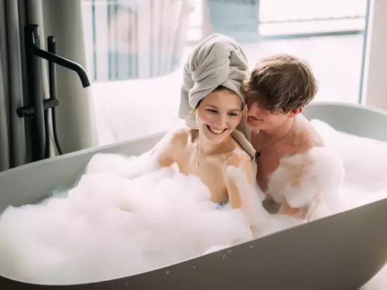 Ein Paar sitzt in einer Badewanne voller Schaum. Die Frau hat ein Handtuch auf dem Kopf und lächelt. Der Mann ist nah bei ihr und schaut sie liebevoll an. Der Raum wirkt hell und freundlich mit einem großen Fenster im Hintergrund.