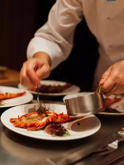 Ein Koch richtet ein Gericht an, das aus einer gebratenen Garnele auf einem weißen Teller besteht. Mit einer kleinen Kelle fügt er Sauce und Garnitur hinzu. Mehrere ähnliche Teller sind im Hintergrund sichtbar.