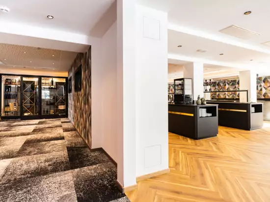 Der Bildinhalt zeigt einen modernen Empfangsbereich mit Holzfußboden und stilvollen Möbeln. Auf der linken Seite sind Regale mit Weinflaschen sowie ein Fernseher zu sehen. Die Wände sind in einem geometrischen Muster gestaltet, während die Beleuchtung den Raum freundlich erhellt.