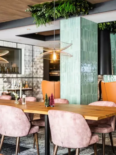 Ein modern eingerichteter Speisesaal mit einem langen Holztisch und bequemen, rosa Stühlen. Auf dem Tisch stehen kleine, dekorative Bäume. Die Atmosphäre ist hell und einladend, mit stilvollen Lampen und modernen Wandfliesen. Pflanzen sind an der Decke angebracht, um eine natürliche Stimmung zu schaffen.