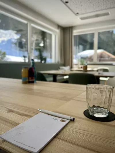 Ein gepolsterter Tisch mit hellem Holz, darauf liegen ein Notizblock und ein Stift. Daneben steht ein Glas auf einem Untersetzer. Im Hintergrund sind große Fenster, durch die Licht strömt, und man sieht eine grüne Umgebung.