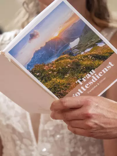 Ein Brautpaar hält ein Programmheft für den Traugottesdienst in der Hand. Das Cover zeigt eine malerische Landschaft mit Bergen und einem Sonnenuntergang. Der Titel "Traugottesdienst" und die Namen "Franziska & Oliver" sind deutlich sichtbar.