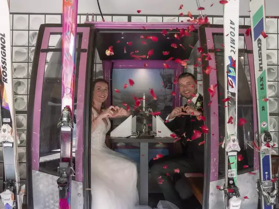 Ein Brautpaar sitzt in einer Gondel und bildet mit ihren Händen ein Herz. Rote Rosenblüten fallen um sie herum. Die Braut trägt ein weißes Kleid, der Bräutigam einen schwarzen Anzug. Im Hintergrund ist eine festliche Tischdeko zu sehen.