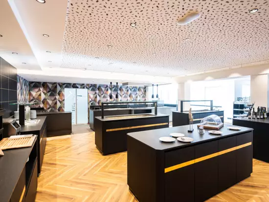 Die Bildbeschreibung zeigt eine moderne Restaurantküche mit schwarzen Möbeln und eleganten Einrichtungen. Der Boden ist aus Holz, und die Wände sind farblich gestaltet. Auf den Tischen liegen Teller und Besteck, und es gibt eine Vitrine mit Getränken im Hintergrund.