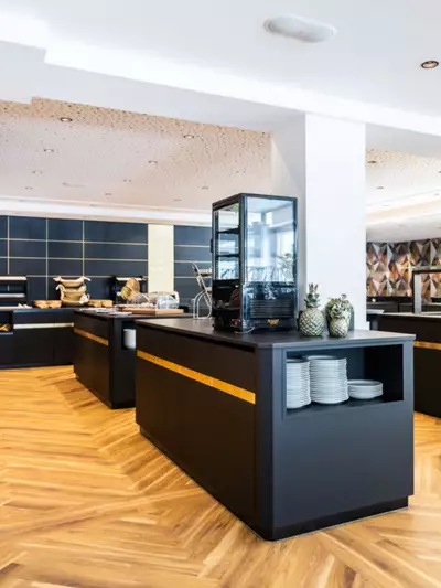 Moderne Buffet-Umgebung mit eleganten, schwarzen Verkaufstheken und Holzfußboden. Auf den Theken sind verschiedene Lebensmittel und Getränke platziert. Ein Bereich ist durch Glas abgegrenzt, und frische Ananas sind dekorativ aufgestellt. Die Lichtverhältnisse sind hell und einladend.