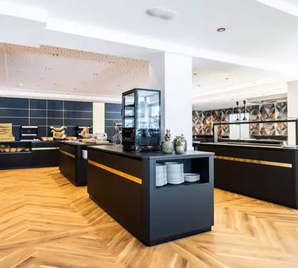 Moderne Buffet-Umgebung mit eleganten, schwarzen Verkaufstheken und Holzfußboden. Auf den Theken sind verschiedene Lebensmittel und Getränke platziert. Ein Bereich ist durch Glas abgegrenzt, und frische Ananas sind dekorativ aufgestellt. Die Lichtverhältnisse sind hell und einladend.