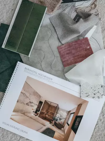 Eine Ansammlung von verschiedenen Stoffmustern und Farben liegt um ein Heft, das die Innenarchitektur einer Saunawohlfühlsuite zeigt. Die Abbildung zeigt ein modernes, elegantes Schlafzimmer mit Holz- und Steinelementen. Der Titel des Hefts lautet „Berghotel Hochfügen“.