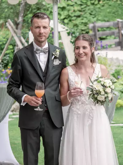 Ein Brautpaar steht in einem Garten. Der Bräutigam trägt einen dunklen Anzug mit einer Fliege und hält ein Glas mit einem orangen Getränk. Die Braut in einem eleganten, hellen Kleid hält einen Sektglas und einen Blumenstrauß mit weißen Blumen. Beide lächeln glücklich.