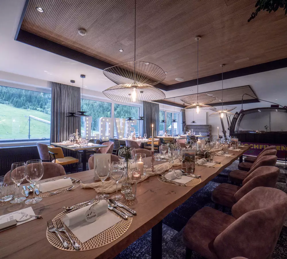 Ein eleganter Speisebereich mit einem langen Tisch, festlich eingedeckt mit Gläsern und Besteck. Sanftes Licht strömt durch große Fenster mit Blick auf grüne Hügel. Gemütliche Stühle und dekorative Elemente schaffen eine einladende Atmosphäre für ein besonderes Essen.