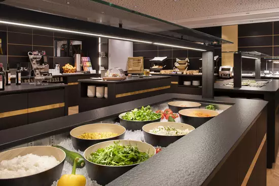 Eine moderne Küchenlandschaft mit schwarzen Möbeln. In großen Schalen sind verschiedene frische Zutaten wie Gemüse, Reis und Salat auf Eis präsentiert. Im Hintergrund sind Küchengeräte und eine Auswahl an Getränken sichtbar. Die Atmosphäre ist hell und einladend.