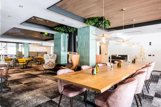 Ein modernes Restaurant mit einem langen Holztisch in der Mitte, umgeben von gepolsterten Stühlen in sanften Farben. Grünes Blattwerk hängt von der Decke, und auf dem Tisch stehen kleine dekorative Bäume. Die Atmosphäre ist freundlich und einladend.